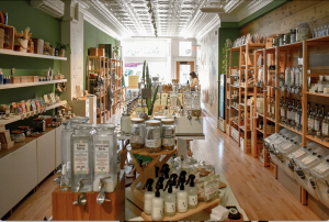 The inside of Green Life Trading Co. storefront. Refill stations for cleaning supplies, spray bottles and soaps in the foreground. A person browses in the background.